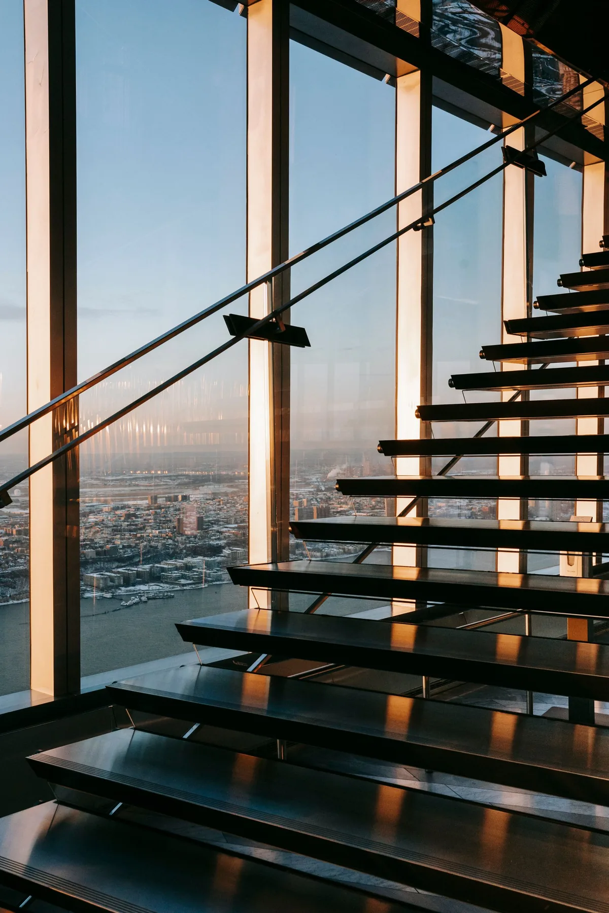 A sunlit staircase rising beside glass walls, with a wide view across the city.